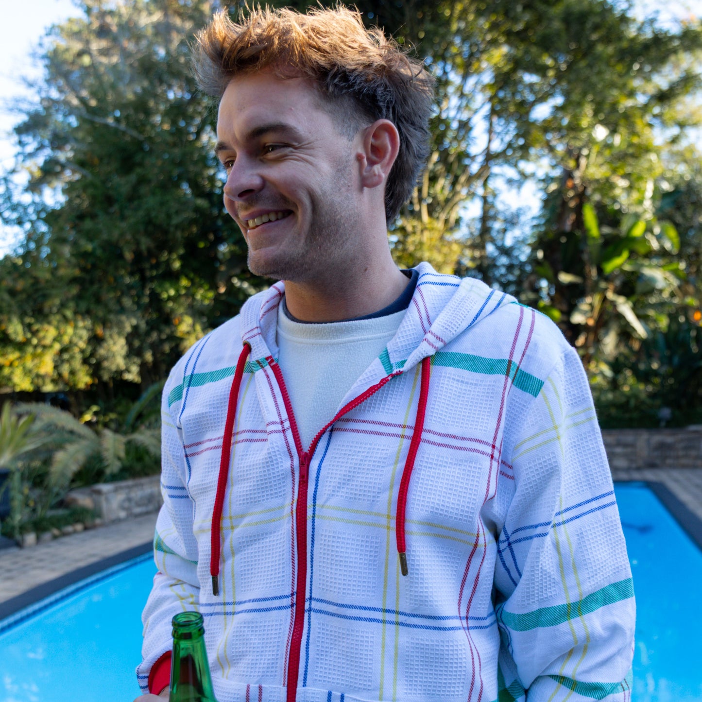 Man holding a beer by a pool with trees in the background, wearing a hoodie made from dishcloth fabric