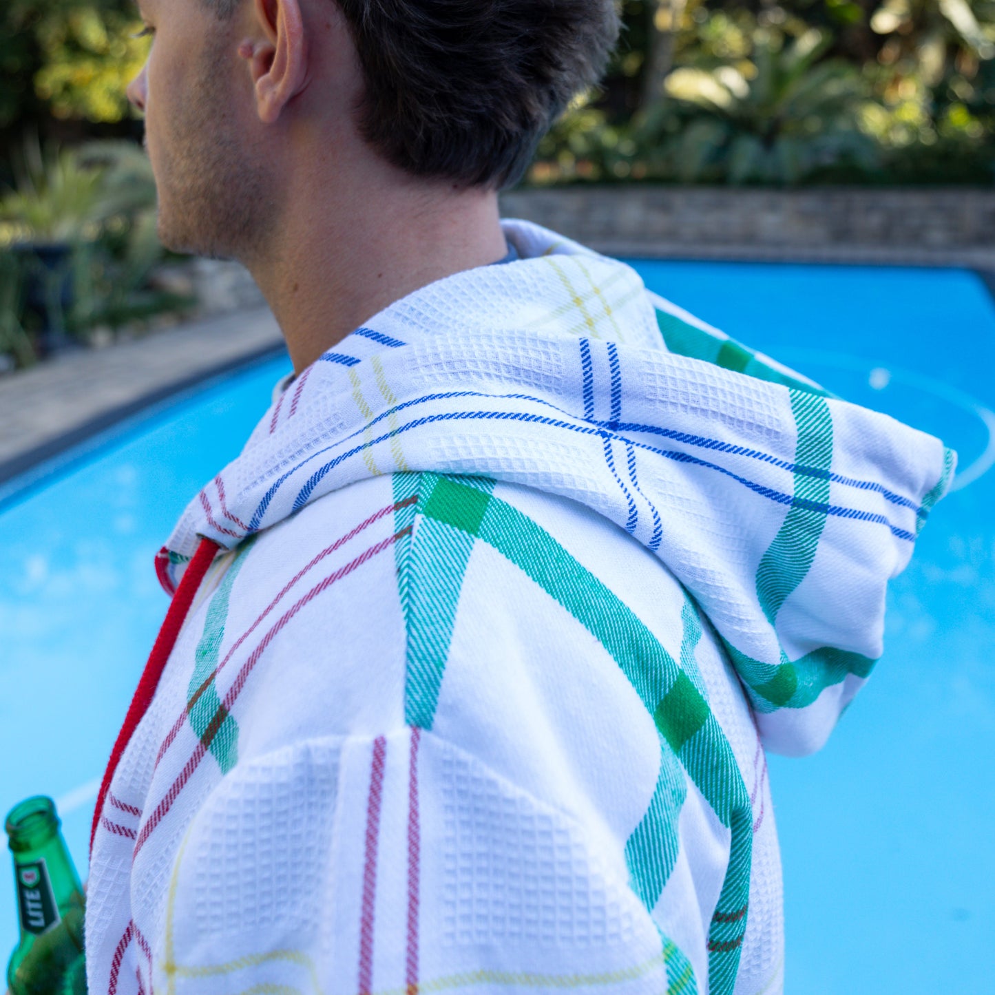 Man wearing a hoodie made from dishcloth fabric, standing next to a pool, with a beer in hand