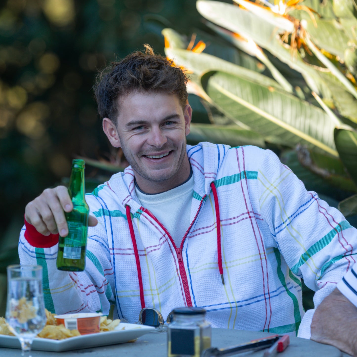 Man holding a beer bottle outdoors with greenery in the background, wearing a hoodie made from dishcloth fabric